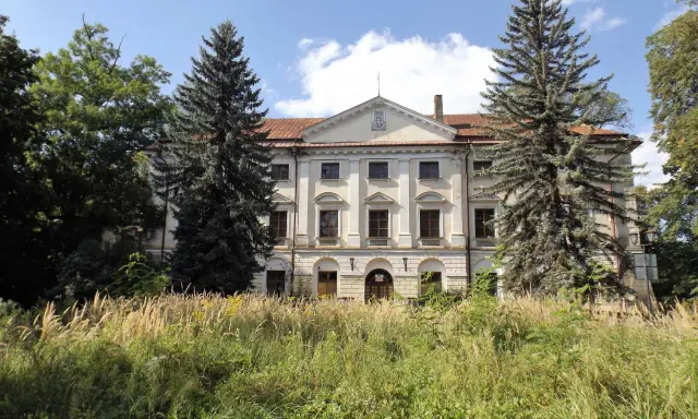 Dawny dwór otoczony przez wysokie trawy i drzewa, w tym sosny i dęby.