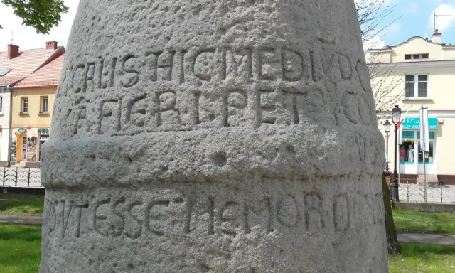 Kamienny słup milowy z wyrytymi łacińskimi napisami, stojący na tle budynków i zieleni.