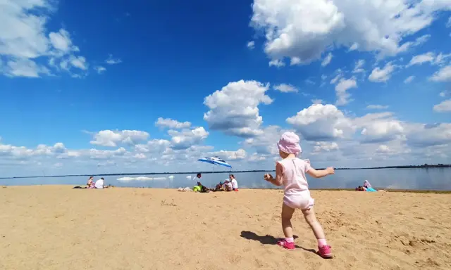 Dziewczynka w różowym kapelusiku bawi się na plaży przy zaporze Jeziorsko. W tle widać ludzi odpoczywających pod parasolem i spokojną taflę wody.