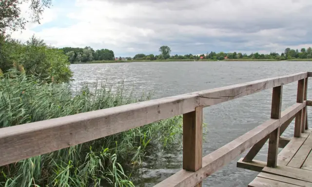 Drewniana kładka nad spokojnym jeziorem Lednica, otoczona trzcinami i zielenią.