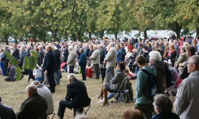 Tłum ludzi zgromadzony na trawie, drzewa w tle. Odpust w Bieniszewie przyciągnął wielu wiernych.