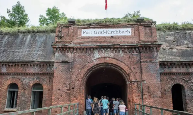 Wejście do Fortu Graf-Kirchbach, historycznej twierdzy w Poznaniu. Grupa turystów wchodzi do ciemnego tunelu.