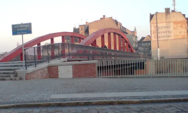 Czerwony most Biskupa Jordana z kłódkami na balustradach, otoczony kamienicami.