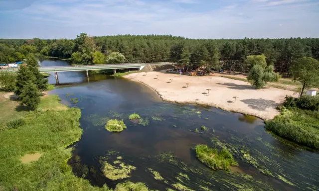 Malownicza plaża nad jedną z wielkopolskich rzek, z mostem i lasem w tle. Idealne miejsce na wypoczynek.