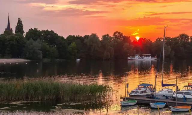 Zachód słońca nad jeziorem z piaszczystą plażą w Polsce. Łodzie zacumowane przy pomoście, na wodzie pływają łabędzie.