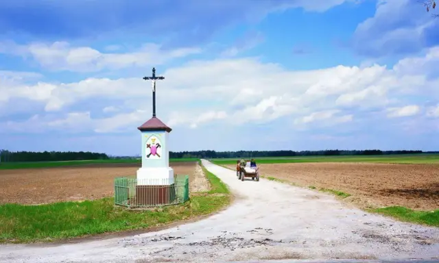 Krzyż przy drodze, obok pola i lasu. Na drodze widać wóz ciągnięty przez konie, z ludźmi.