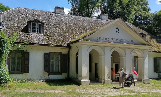 Stare dworki polskie z omszałym dachem i werandą. Rodzina z wózkiem spaceruje po dziedzińcu.