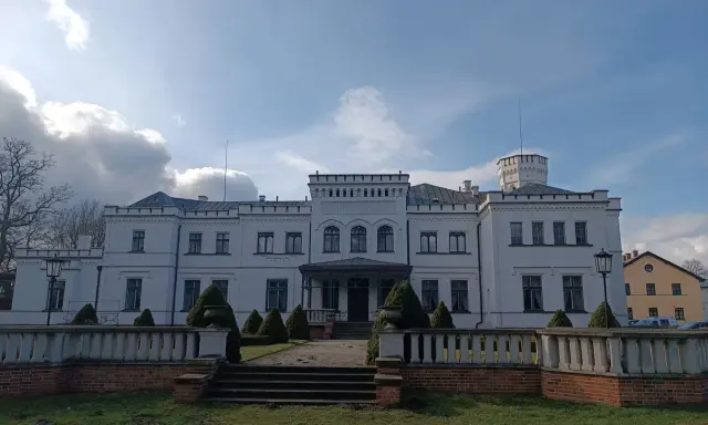 Biały pałac w Będlewie z wieżyczką i ozdobnym murem. Słońce przebija się przez chmury, tworząc malowniczy krajobraz.