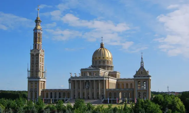 Imponujące sanktuarium koło Konina z wysoką wieżą i złotą kopułą, otoczone zielenią.