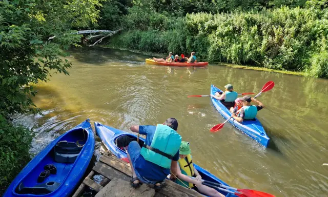 Rodzina pływa kajakami po rzece Prośnie, otoczonej bujną zielenią.