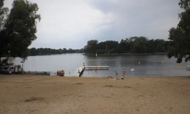 Plaża nad jeziorem koło Gniezna. Ludzie odpoczywają, niektórzy kąpią się. W tle las i spokojna woda.