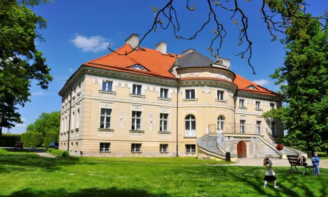 Pałac w Lewkowie, żółty, z pomarańczowym dachem, otoczony zielenią. Dzieci bawią się na trawniku przed budynkiem.