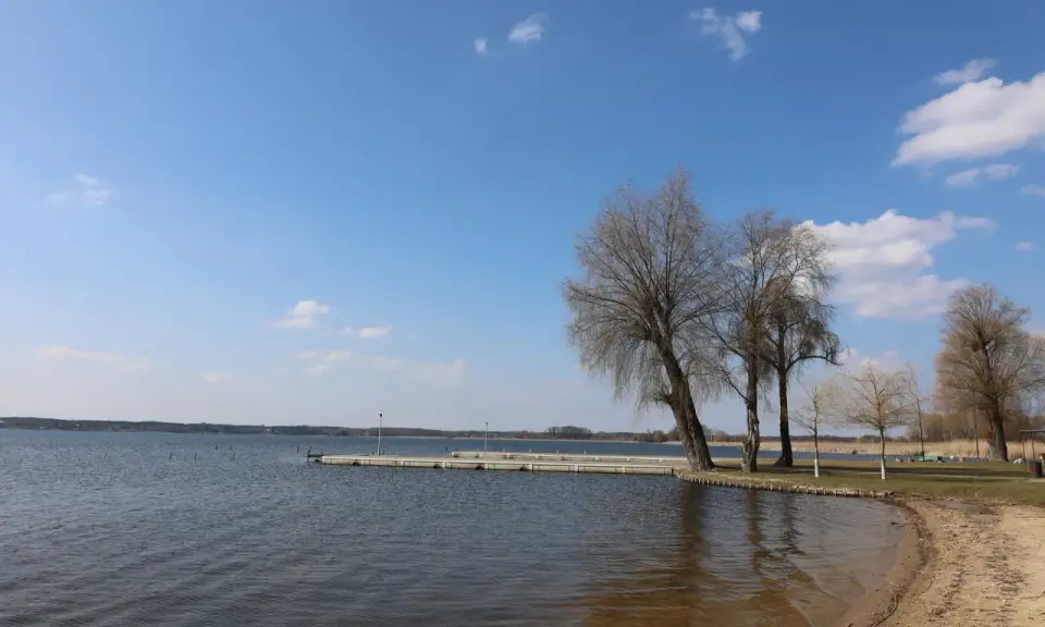 Spokojne jezioro w Zbąszyniu, z piaszczystą plażą i pomostem. Drzewa bez liści tworzą malowniczy krajobraz.