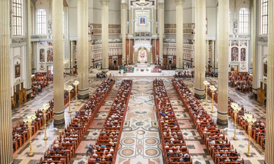Wielka bazylika w Licheniu Nowym wypełniona wiernymi. Długie ławy zapełnione ludźmi, majestatyczne kolumny i ołtarz w tle.