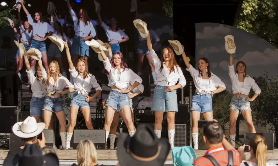 Dziewczęcy zespół country z Wolsztyna w białych koszulach i jeansowych szortach tańczy na scenie, unosząc kapelusze.