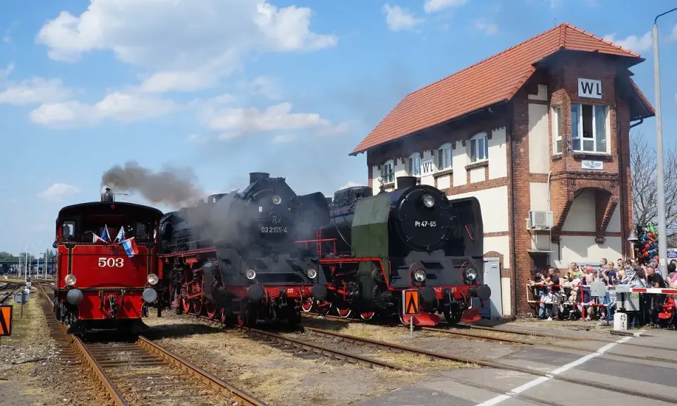 Parada parowozów z parą unoszącą się w powietrzu. Trzy lokomotywy stoją na torach obok ceglanego budynku stacji, a tłum widzów obserwuje wydarzenie.