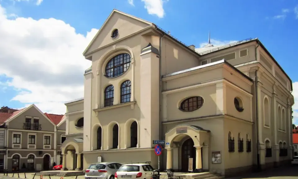 Jasny budynek z charakterystycznymi oknami, jeden z leszczyńskich zabytków, stoi na tle błękitnego nieba z chmurami.
