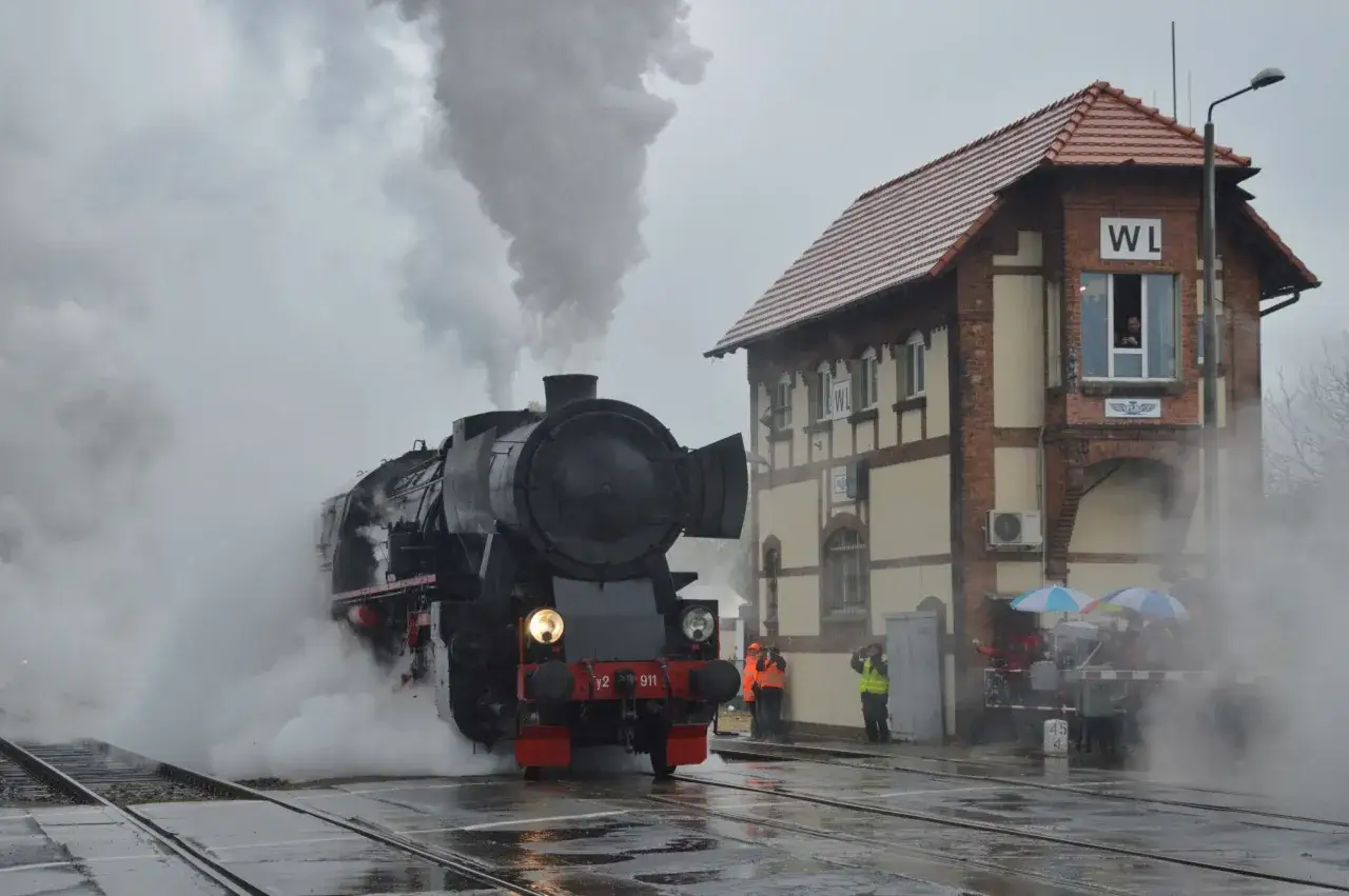 Parada parowozów w deszczu. Potężny, czarny parowóz z numerem 911 wyłania się z mgły i pary, obok zabytkowego budynku stacji kolejowej.