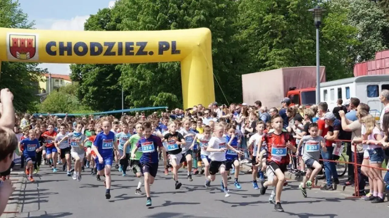 Dzieci biegną w wyścigu pod żółtym łukiem z napisem CHODZIEZ.PL. Wiele radosnych wydarzeń Chodzież czeka na młodych sportowców.