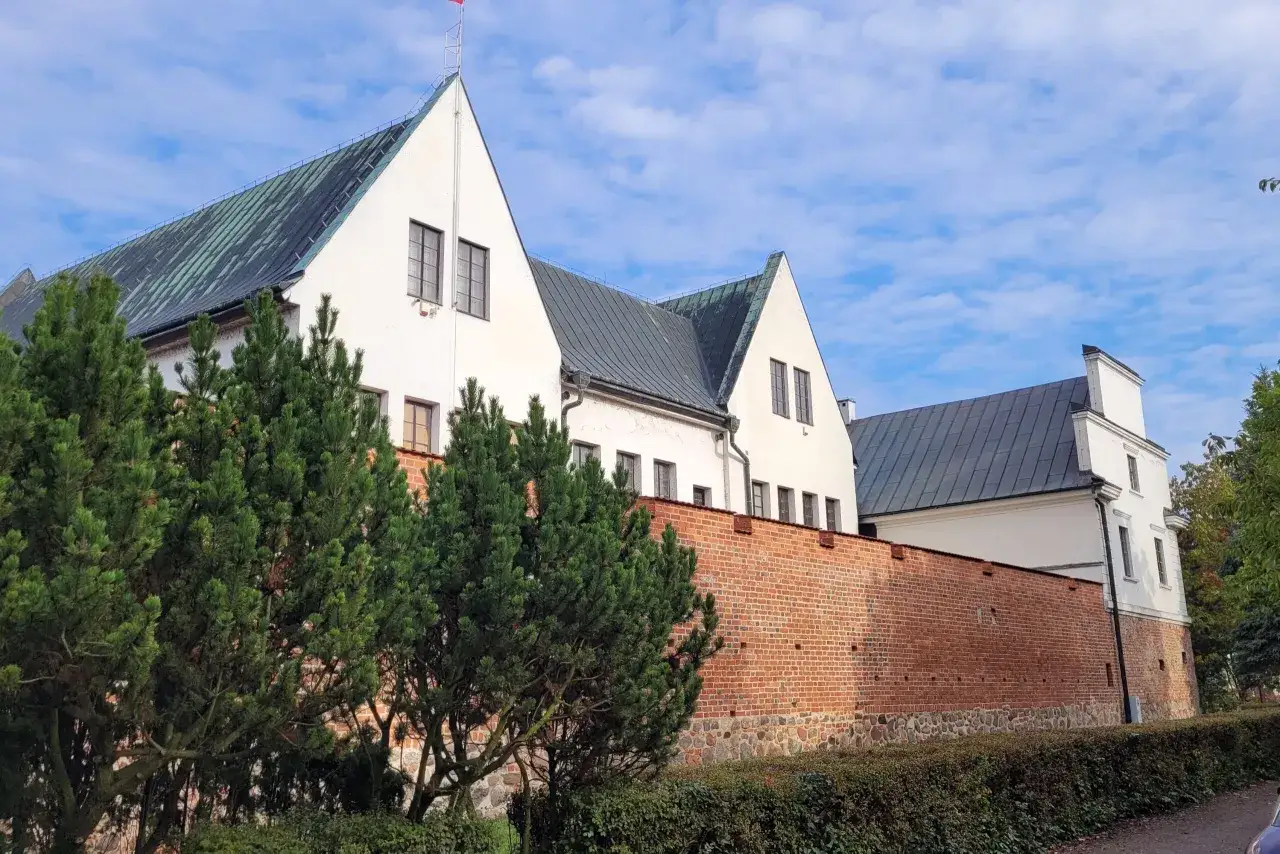 Białe budynki z zielonymi dachami i ceglanym murem obronnym tworzą malowniczy zamek w Koninie.