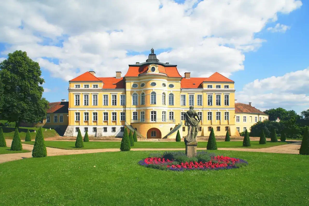 Ż&oacute;łty kurnik pałac z czerwonym dachem, otoczony zielonym ogrodem i rzeźbą.