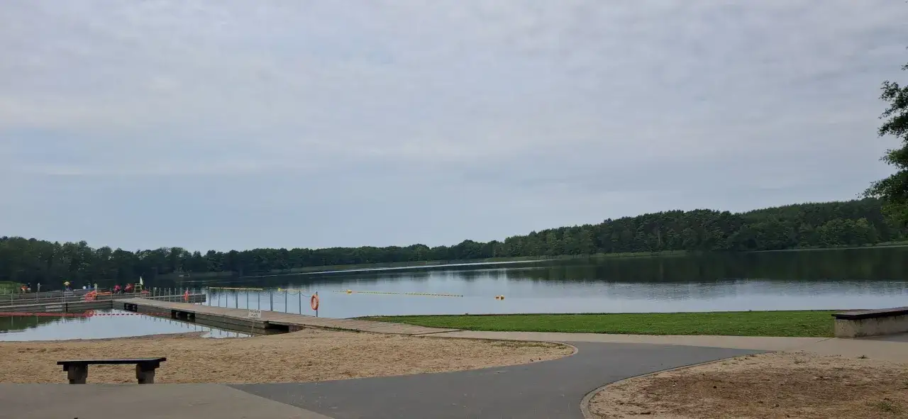 Spokojny dzień nad jeziorem w Puszczy Noteckiej. Drewniane molo, piaszczysta plaża i las odbijający się w tafli wody.