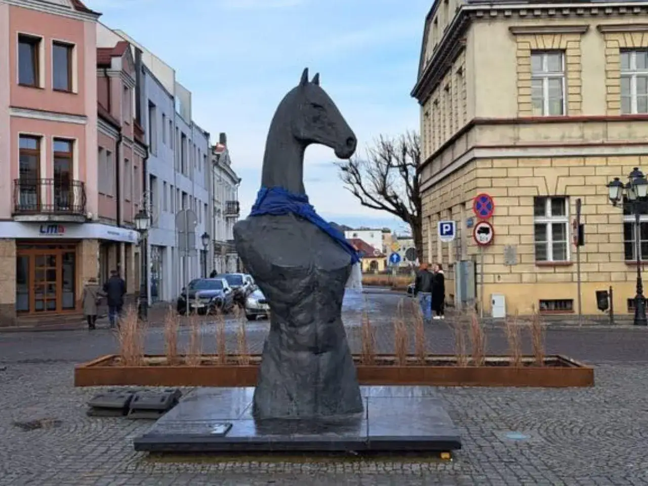 Pomnik konia w Koninie, z niebieską szarfą na szyi, stoi na brukowanym placu. W tle kamienice i drzewa.