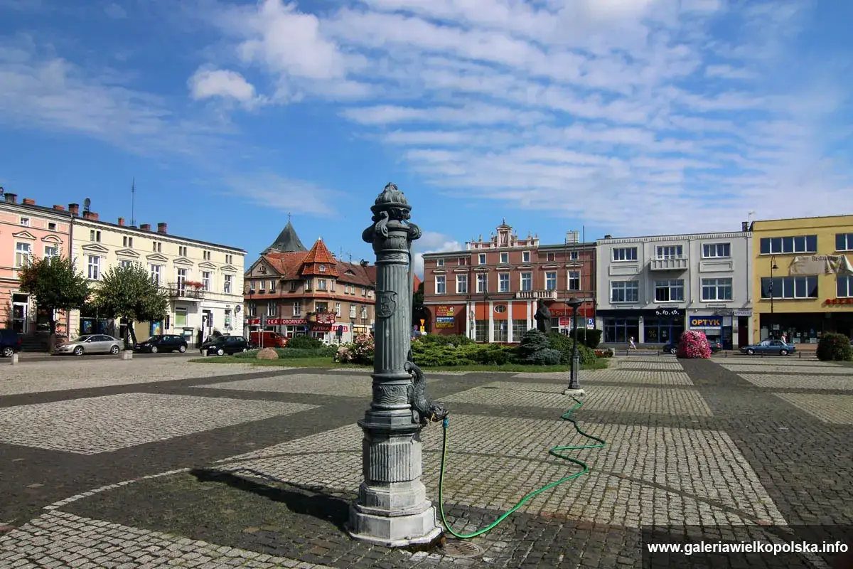 Rynek w Obornikach Wielkopolskich z zabytkową fontanną i kolorowymi kamienicami.