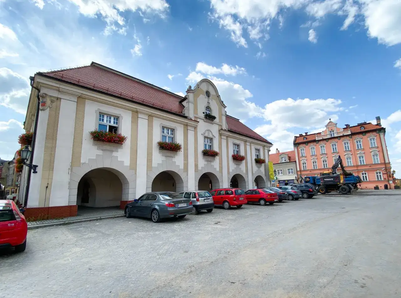 Ratusz w Jarocinie z arkadami i kwiatami w oknach. Na placu zaparkowane samochody i koparka.