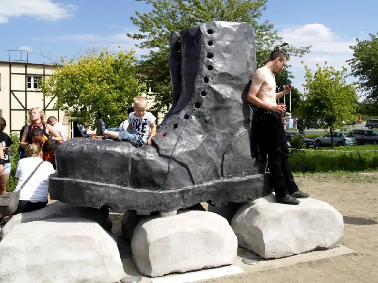 Pomnik glana, gigantycznego buta, przyciąga uwagę. Dziecko siedzi na nim, a młody mężczyzna opiera się o jego bok.