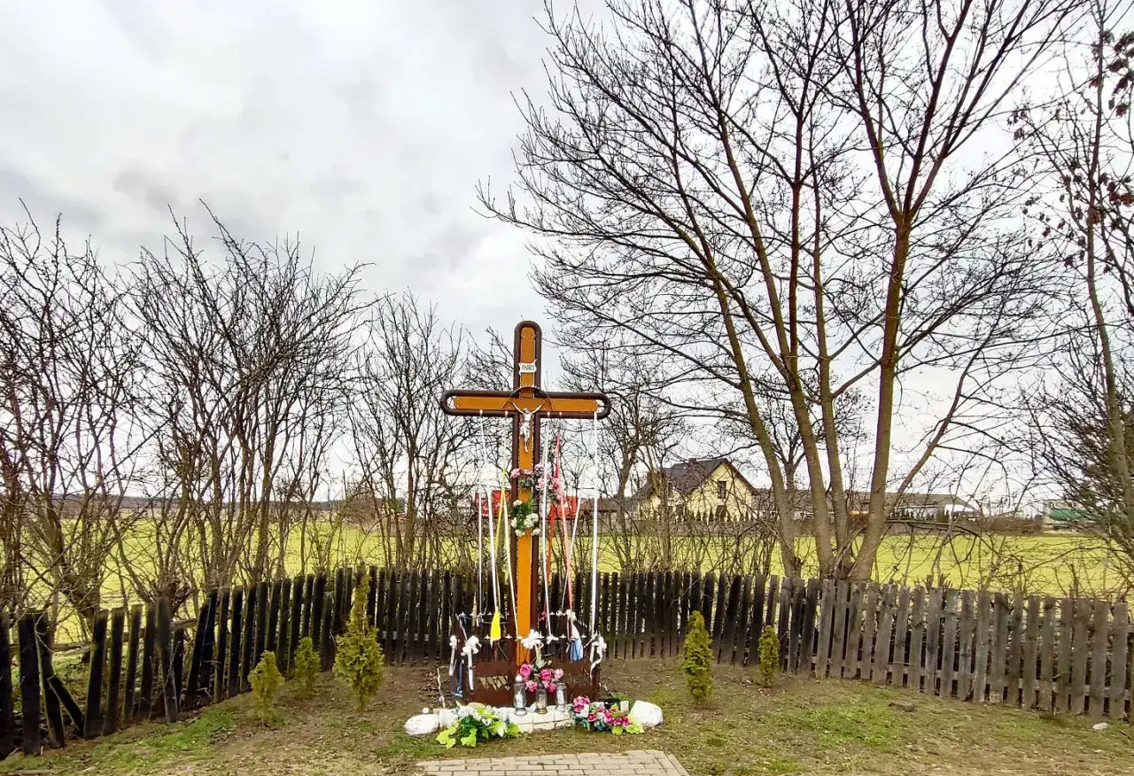 Drewniany krzyż przy drodze, ozdobiony wstążkami i kwiatami, stoi pośród nagich drzew.