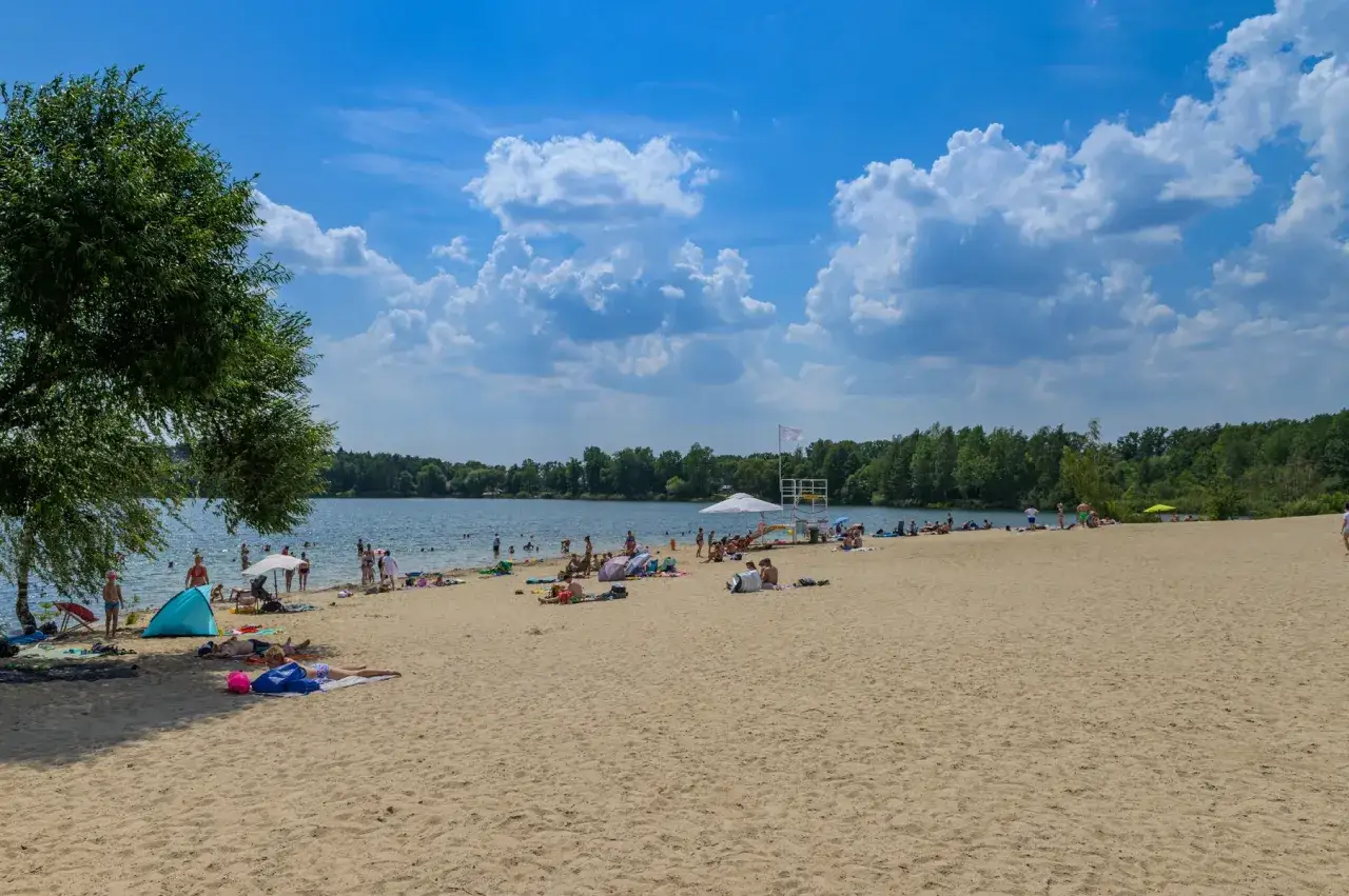Słoneczny dzień nad jeziorem z piaszczystą plażą w Polsce. Ludzie odpoczywają, kąpią się i opalają.