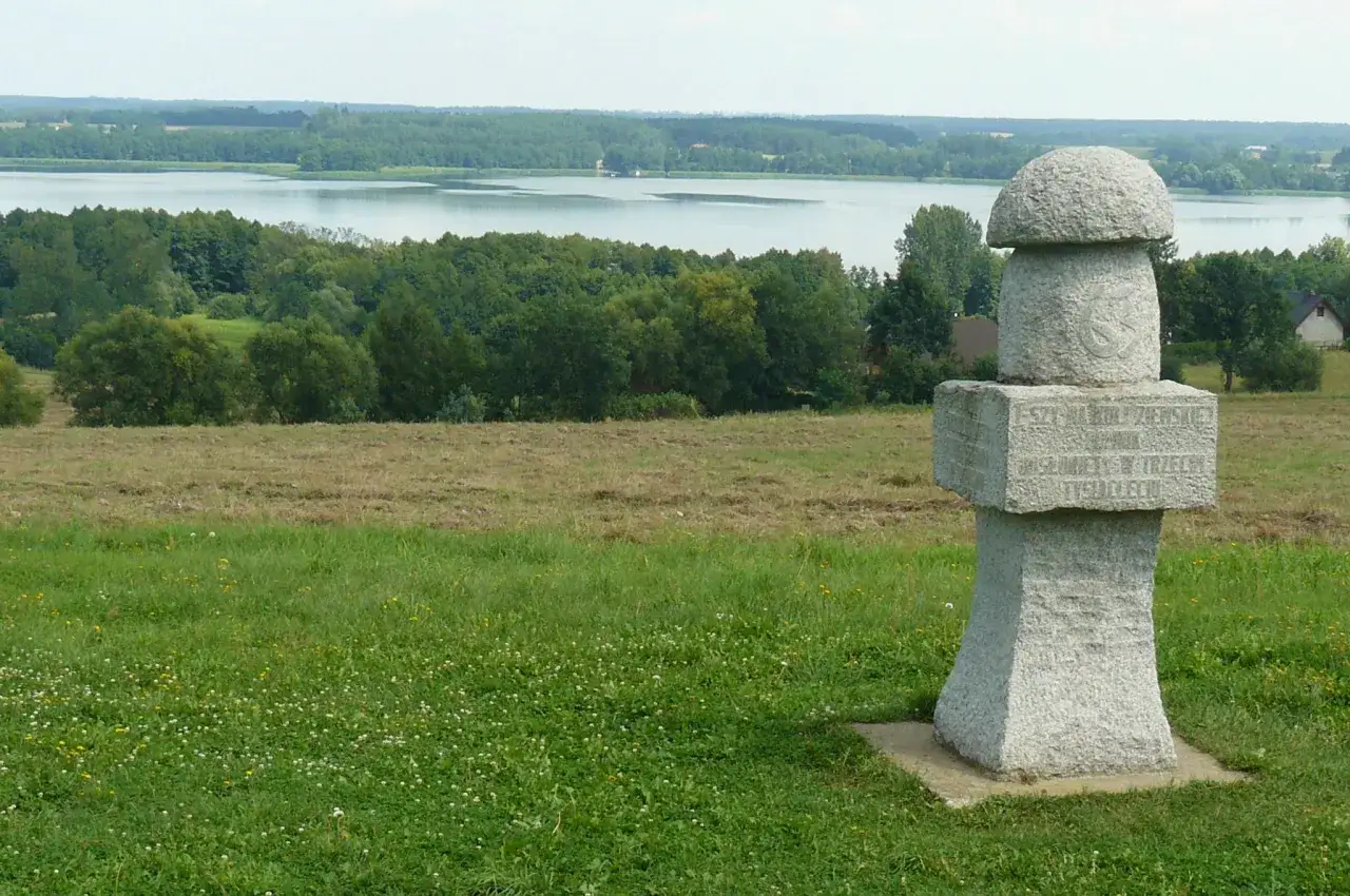 Kamienny, wielki grzyb z inskrypcją stoi na wzgórzu. W tle jezioro i las.