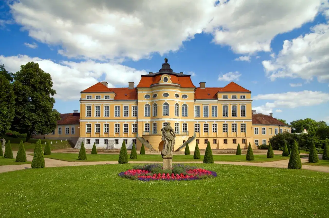 Ż&oacute;łty pałac z czerwonym dachem, otoczony zielonym ogrodem z rzeźbą i kwiecistą rabatą. Piękny rogalin park.