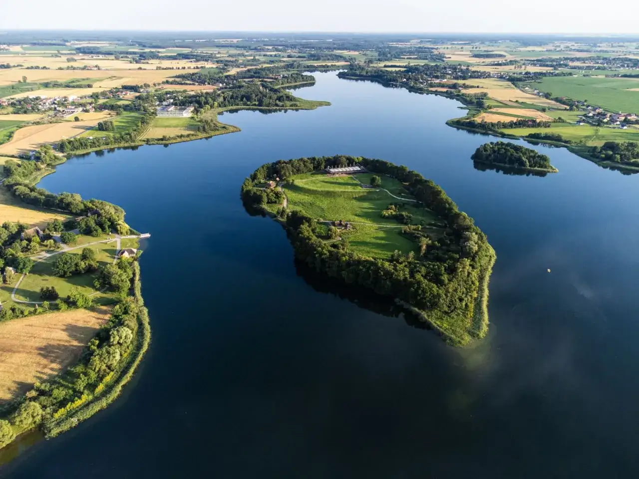Malownicze jezioro Lednica z wyspą Ostr&oacute;w Lednicki, porośniętą drzewami i zielenią, z widocznymi budynkami. W oddali pola i zabudowania.