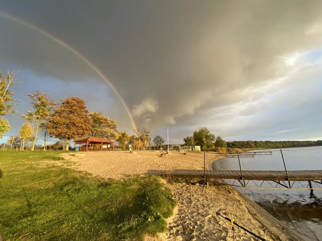 Tęczowy łuk nad piaszczystą plażą, gdzie nad jezioro w Wielkopolsce zaprasza drewniany pomost.