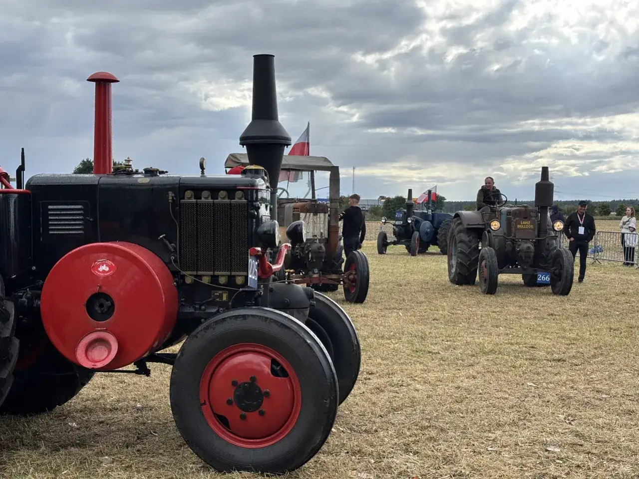 Na festiwalu starych ciągnik&oacute;w i maszyn rolniczych widać czarny traktor z czerwonymi detalami, obok inne zabytkowe maszyny i ludzie.