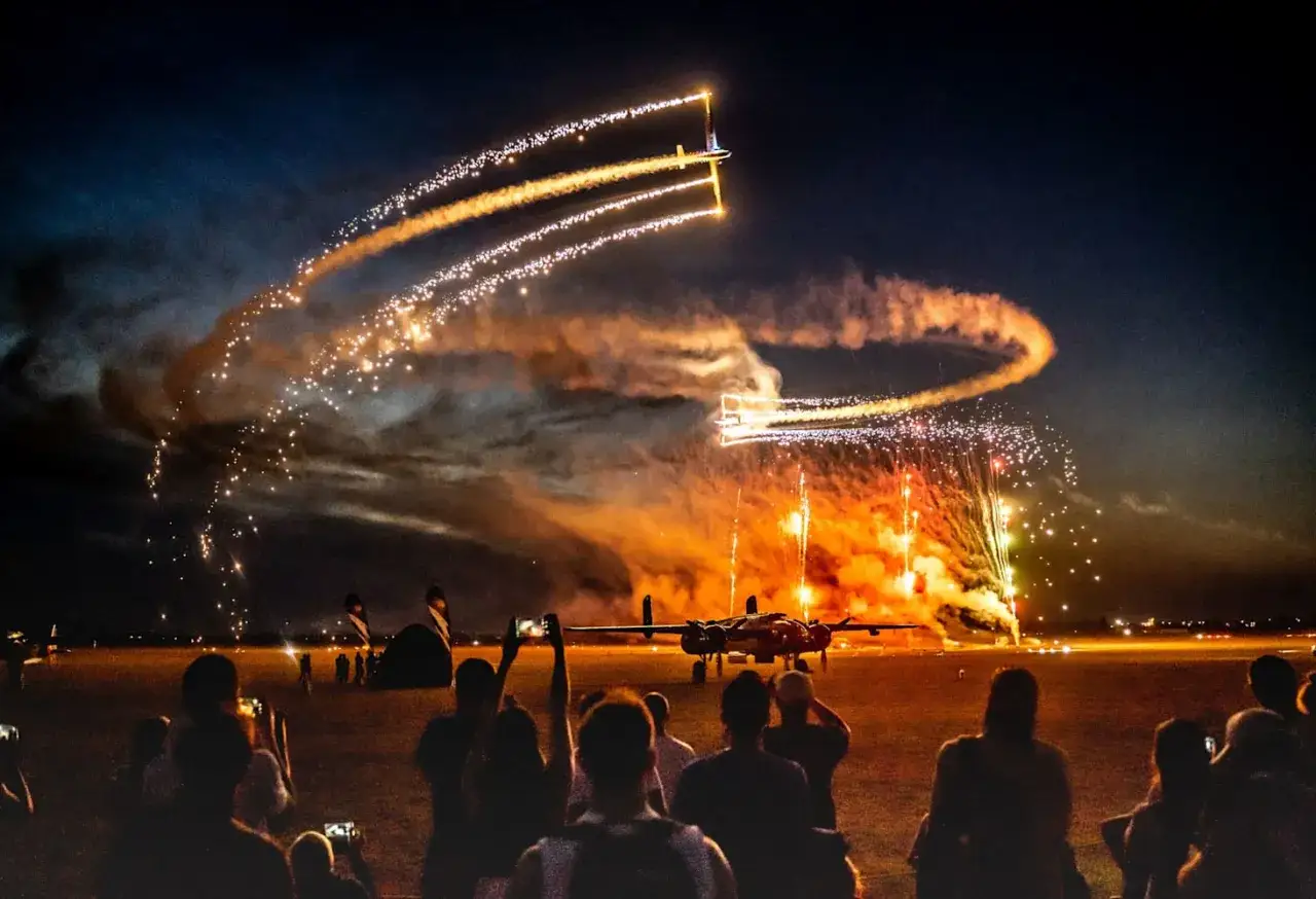Nocne pokazy lotnicze na leszno air show. Samoloty kreślą świetlne ślady, a fajerwerki rozświetlają niebo. Widzowie filmują spektakl.