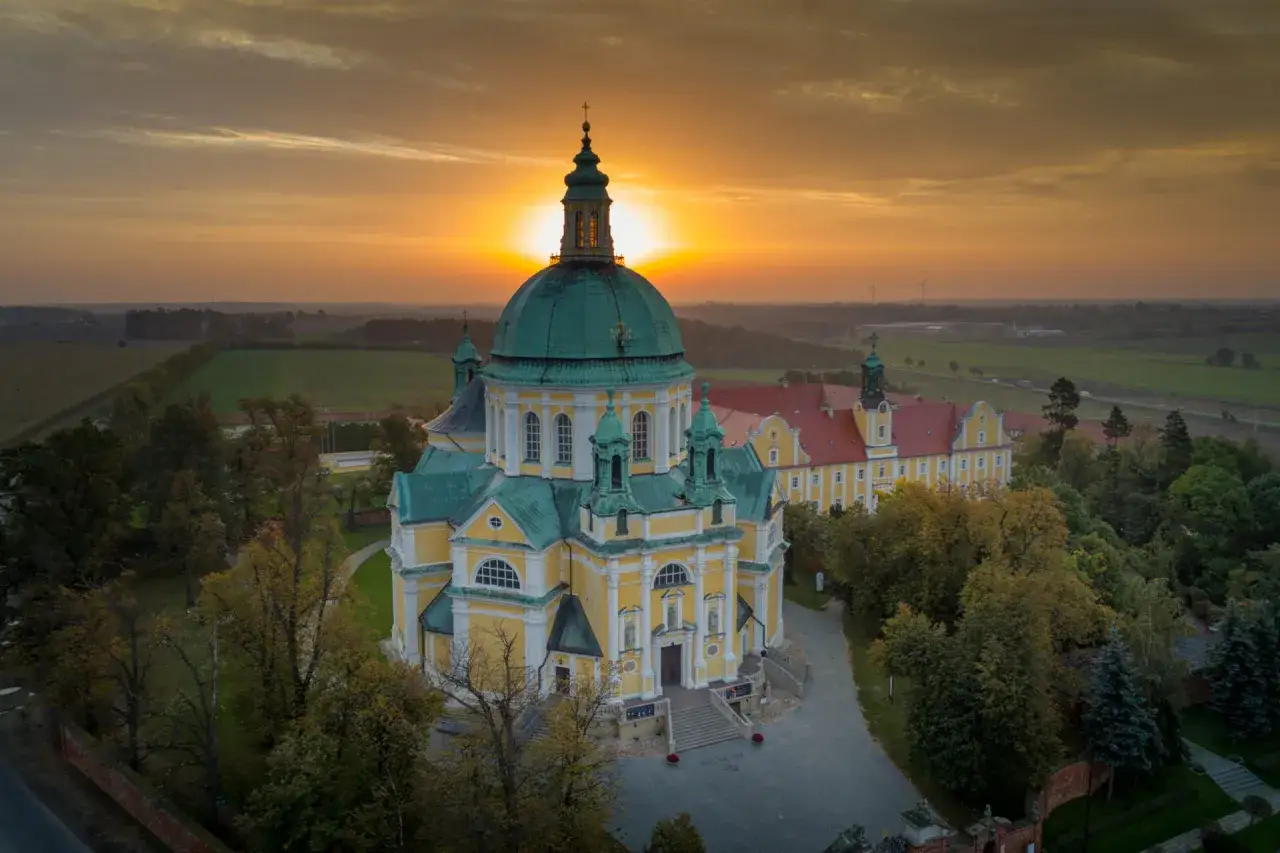 Zachód słońca nad bazyliką na Świętej Górze Gostyń. Majestatyczna budowla z zielonymi kopułami i żółtymi ścianami w otoczeniu drzew.