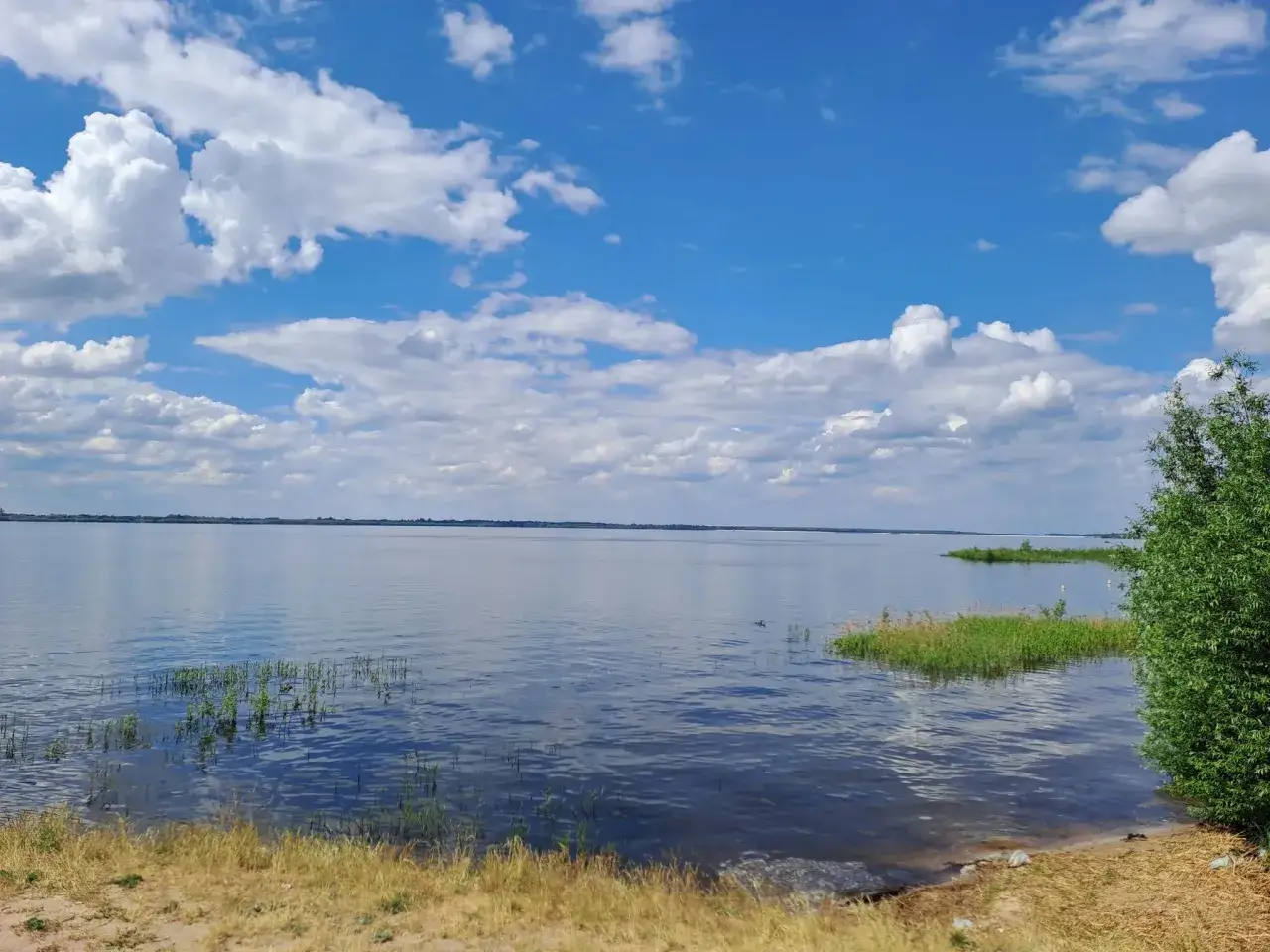 Spokojny dzień nad Jeziorem Jeziorsko. Błękitne niebo z białymi chmurami, spokojna tafla wody i zielone brzegi.
