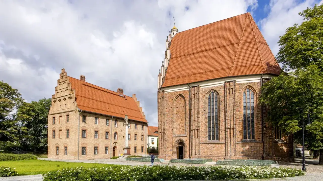 Ostrów Tumski Poznań: ceglane budynki z czerwonymi dachami, katedra i dom kanoników, z zielenią i niebem w tle.
