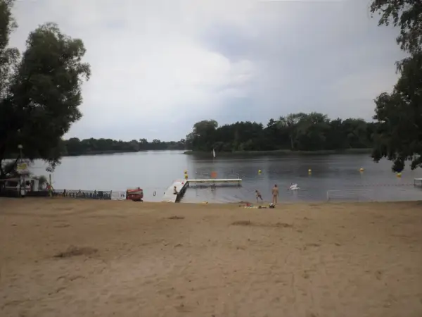Plaża nad jeziorem koło Gniezna. Ludzie odpoczywają, niektórzy kąpią się. W tle las i spokojna woda.