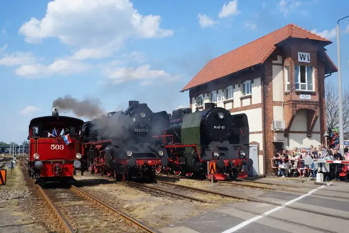 Parada parowozów z parą unoszącą się w powietrzu. Trzy lokomotywy stoją na torach obok ceglanego budynku stacji, a tłum widzów obserwuje wydarzenie.