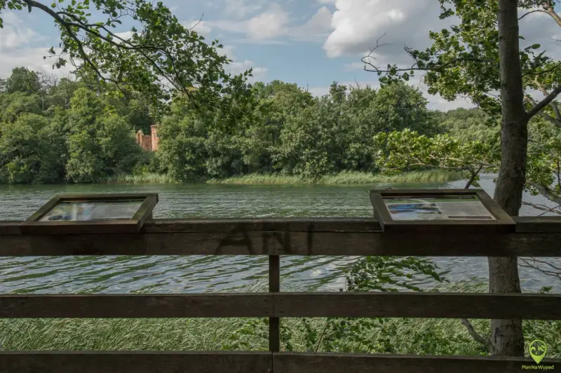 Widok na jezioro w Wielkopolskim Parku Narodowym. Na pierwszym planie drewniana balustrada z tablicami informacyjnymi. W oddali las i ruiny ceglanej budowli.