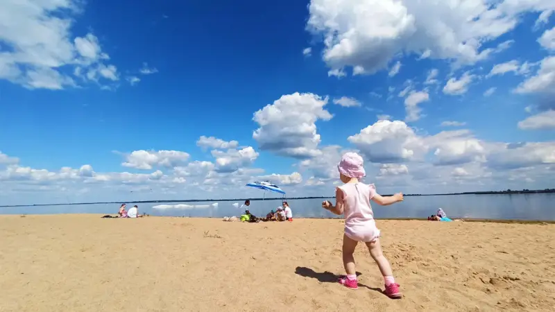 Dziewczynka w różowym kapelusiku bawi się na plaży przy zaporze Jeziorsko. W tle widać ludzi odpoczywających pod parasolem i spokojną taflę wody.